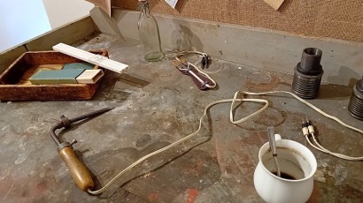 Při pohledu na tento autentický pracovní stůl (ponk) se svěrákem se člověk rázem vrátí tak o 60 let zpět. Nechybí dokonce ani hrnek od kávy se zbytky logru a v pootevřeném šuplíku svačina v promaštěném papírovém sáčku. Na věšáku tehdejší pracovní pláště. 